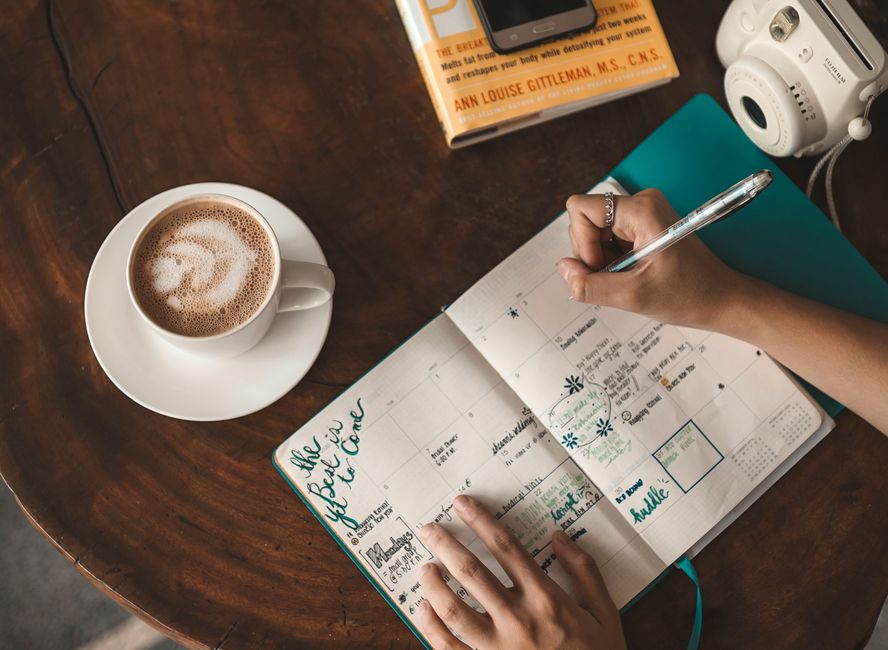 Woman writing in planner with cappuccino, book, and camera on wooden table.